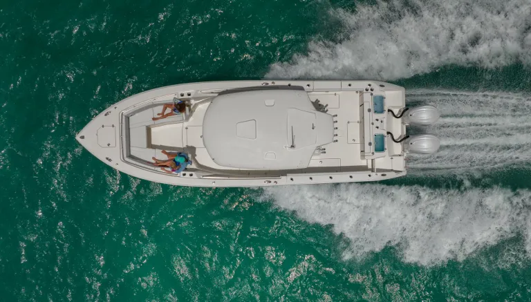 Overhead drone shot of Cobia 320 CC with two women on bow lounge running over green coastal water